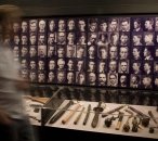 Image of a man moving past a wall covered in early 20th century mugshots. Underneath is a showcase containing weapons.