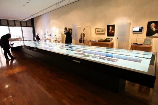 View of a large horizontal backlit display in the Museum of Sydney. Visitors can be seen reading the text and reproductions of Governor Phillip and Governor Macquarie can be seen in the background.