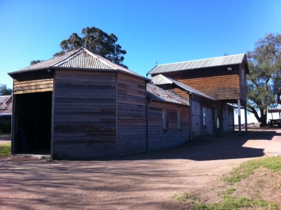 A wooden smokehouse with pointed roof, part of a larger farm complex.